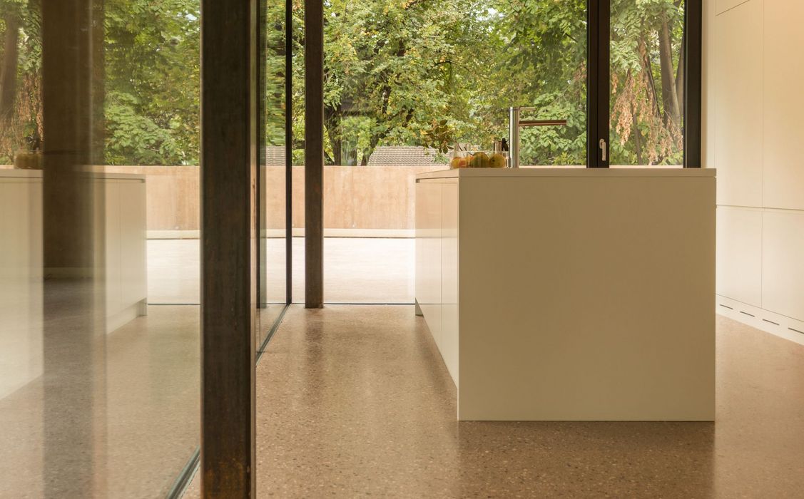 Blick durch eine bodentiefe Glasfront in eine moderne, minimalistische Küche im Erdgeschoss mit einer weißen Kücheninsel und einem polierten Betonboden.