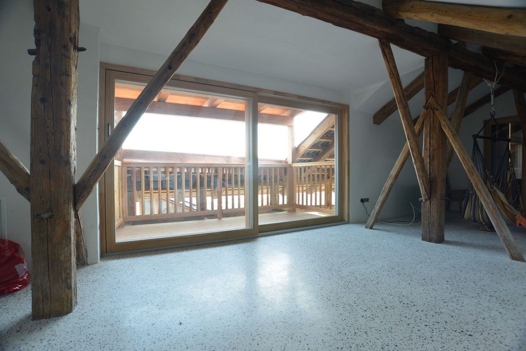 Ein geräumiger Dachboden mit hellem Terrazzoboden und sichtbaren Holzbalken, der durch eine große Schiebetür Blick auf einen hölzernen Balkon bietet.