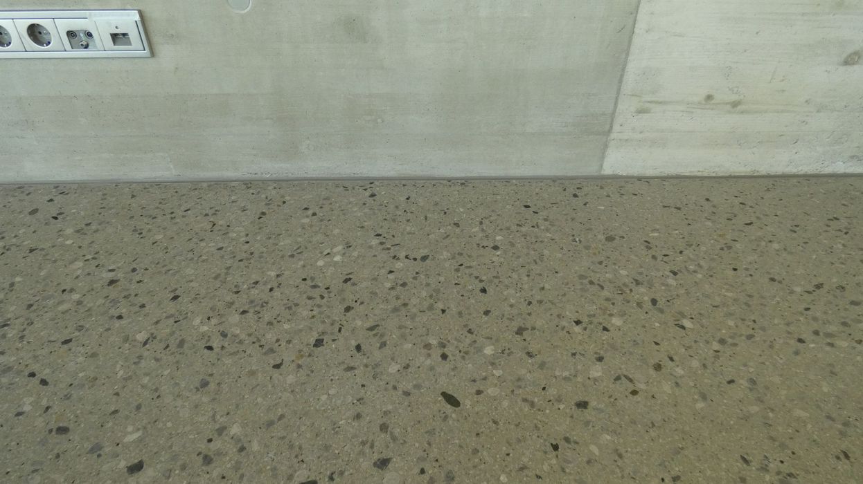 Blick auf einen glatten Terrazzoboden, der an eine schlichte, graue Betonwand angrenzt, an der eine Steckerleiste montiert ist.