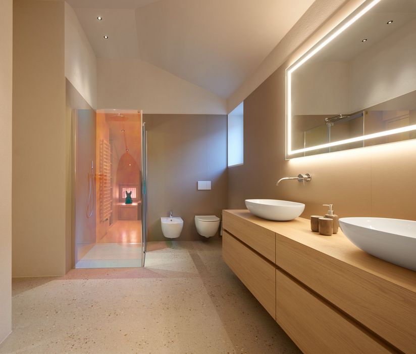 Modernes Badezimmer mit Doppelwaschtisch aus Holz, beleuchtetem Wandspiegel, wandhängendem WC, Bidet und einer gläsernen Dusche mit orangefarbener Beleuchtung.