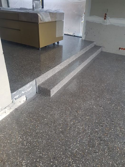 Ein Innenraum in einer Baustelle mit einem frisch verlegten, grauen Terrazzoboden und einer zweistufigen Treppe, die zu einer Küchenzeile führt.