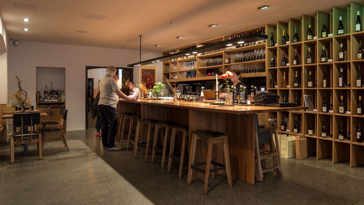 Ein Blick in eine moderne Weinstube mit einem langen Holztresen, an dem Gäste sitzen und eine Bedienung hinter dem Tresen steht, begleitet von einem großen Weinregal an der Wand.