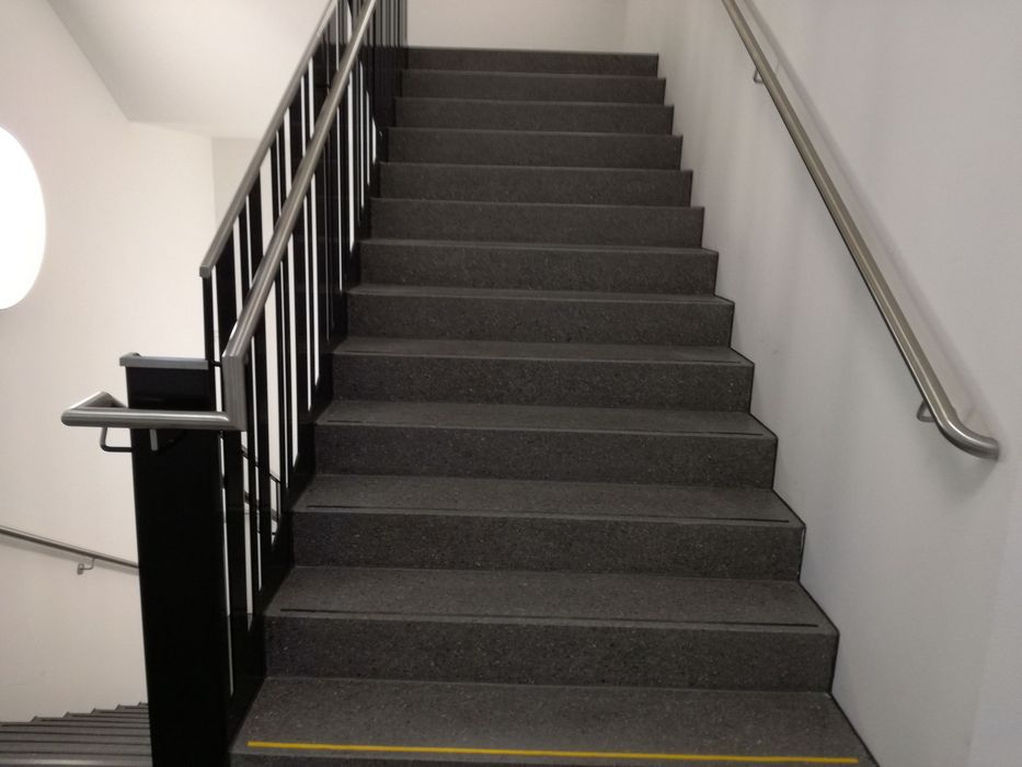 Blick von einer Treppe aus grauem Stein nach oben zu einem weiteren Treppenabsatz, flankiert von einem schwarzen Geländer und einem an der Wand montierten Handlauf aus Metall.