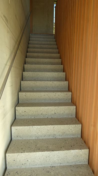 Eine gerade Treppe aus hellgrauem Naturstein führt nach oben, flankiert links von einer Sichtbetonwand mit Handlauf und rechts von einer Wand aus vertikalen Holzlamellen.