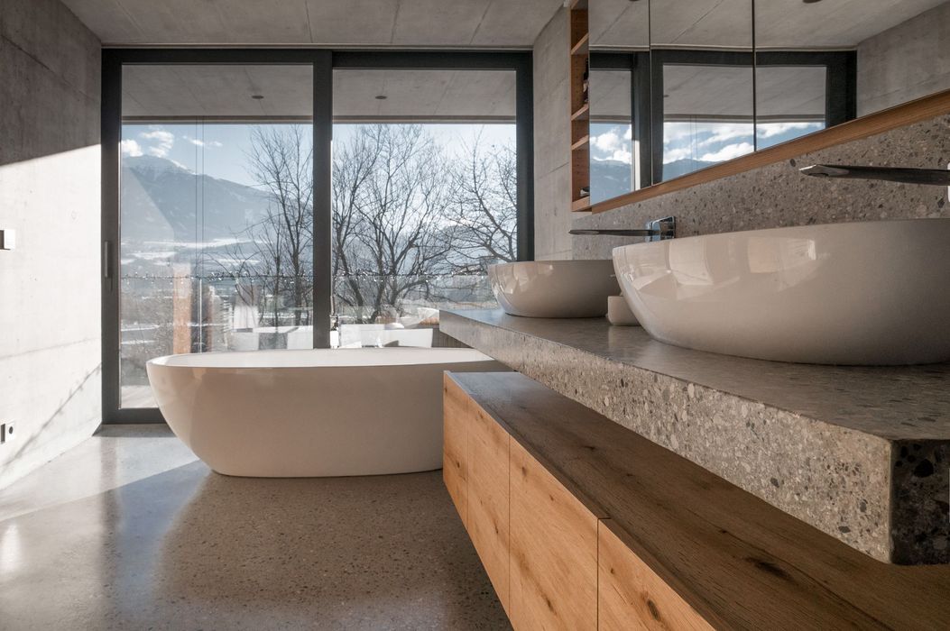 Ein modernes Badezimmer mit zwei weißen Aufsatzwaschbecken auf einer grauen Steinplatte, einem hölzernen Unterschrank, einer freistehenden Badewanne und einer raumhohen Fensterfront mit Blick auf schneebedeckte Berge.
