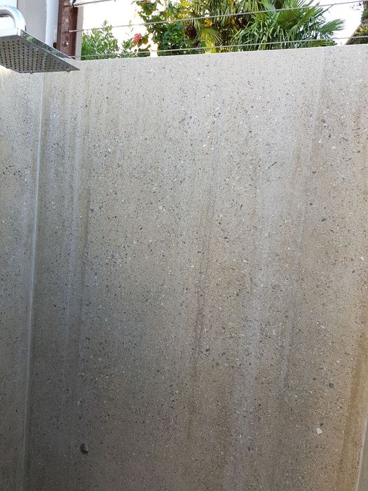 Eine Außendusche mit einem quadratischen Duschkopf vor einer hohen, steinernen Wand, hinter der sich grüne Pflanzen bei Sonnenschein zeigen.