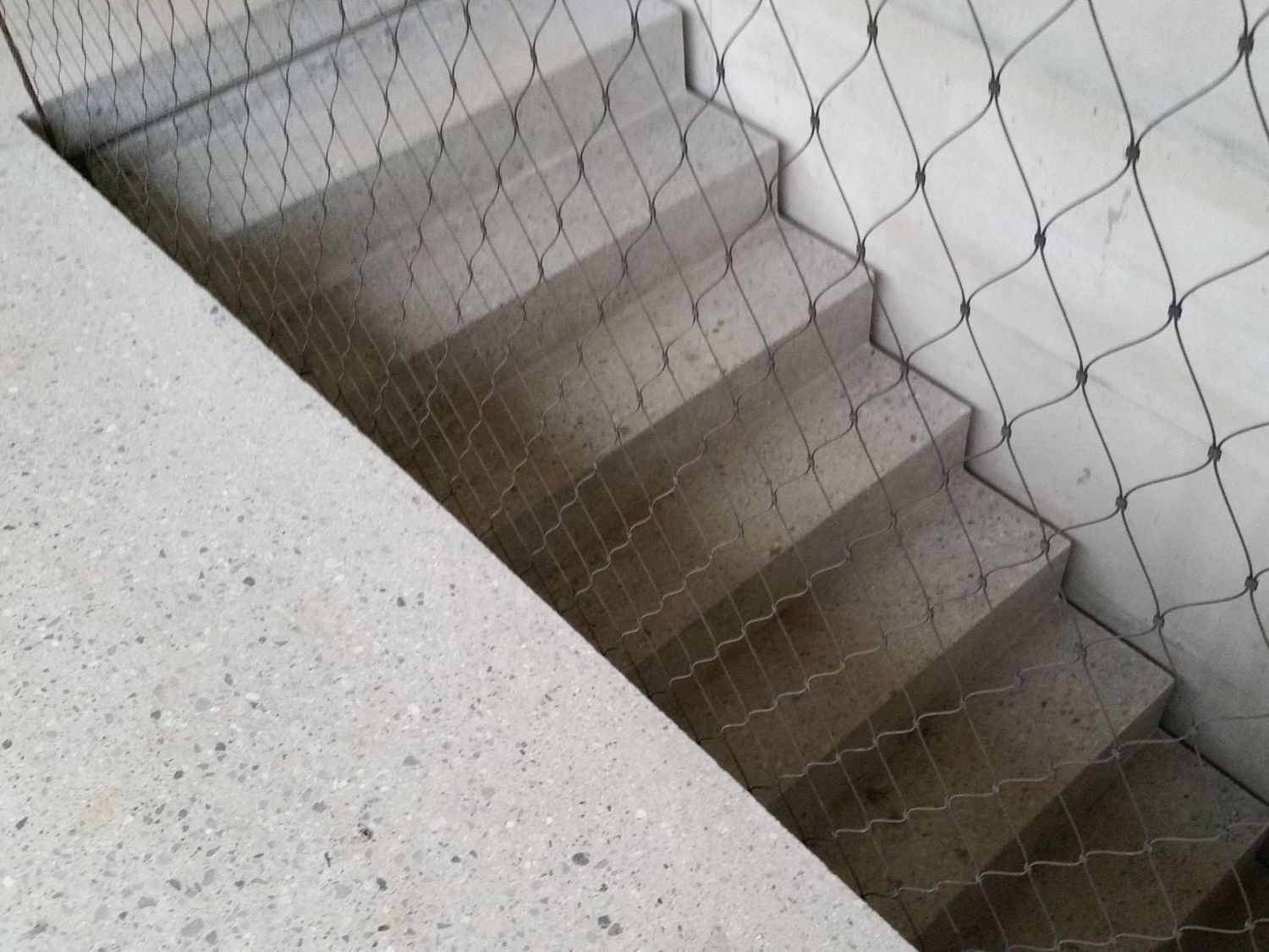 Eine Treppe aus hellem Beton, die hinter einem metallischen Drahtnetz verläuft.