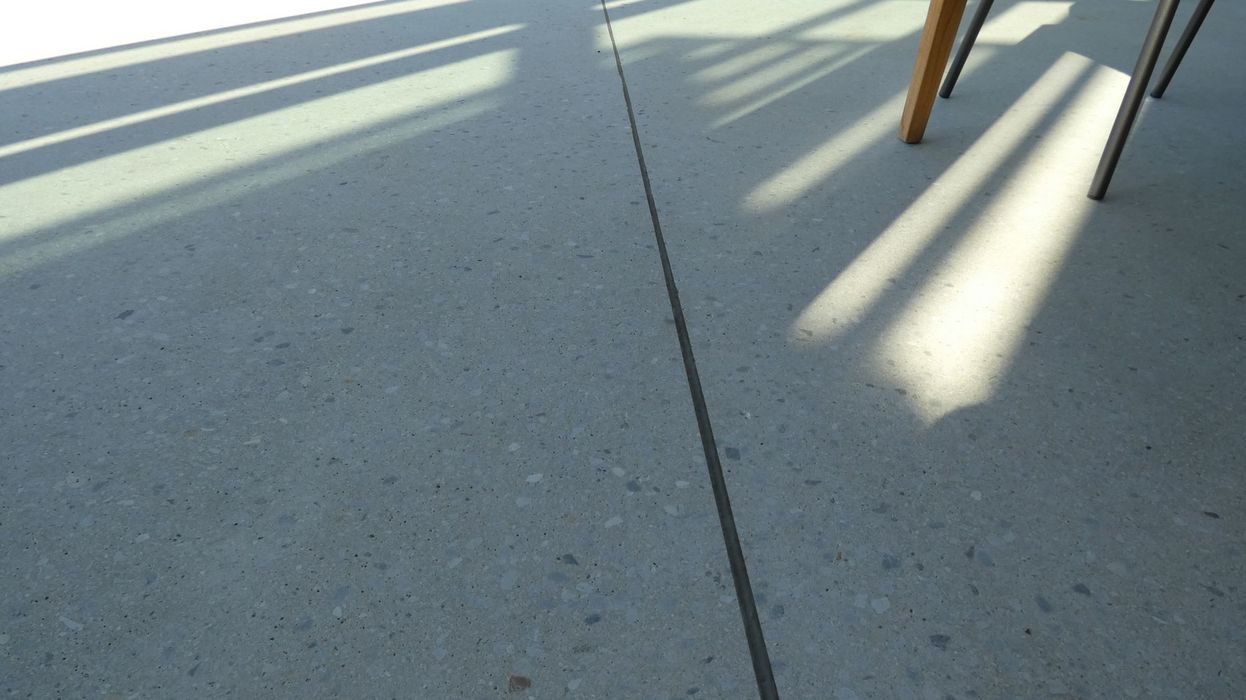 Ein grauer Terrazzo-Boden mit einer durchgehenden Dehnungsfuge, auf den Sonnenlichtmuster von darüber liegenden Strukturen fallen.