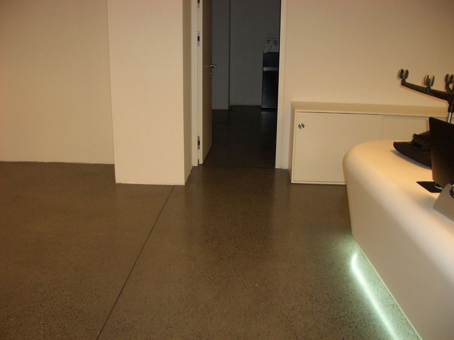 Ein minimalistisch eingerichteter Raum mit einem glatten Betonboden, einer weißen Empfangstheke mit integrierter Unterbeleuchtung und einem geöffneten Durchgang zu einem angrenzenden Raum.