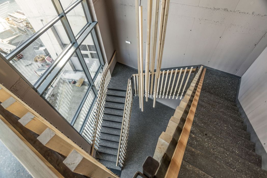 Draufsicht auf ein Treppenhaus mit einer dunkelgrauen Steintreppe, hängenden Holzelementen als Geländer und einer großen Glasfront mit Blick auf eine Baustelle.