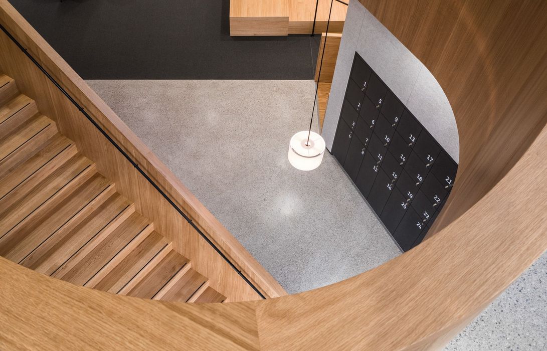 Ein Blick von oben auf eine geschwungene Holztreppe, die in einen Raum mit einer Wand voller nummerierter Schließfächer und einer hängenden Pendelleuchte führt.