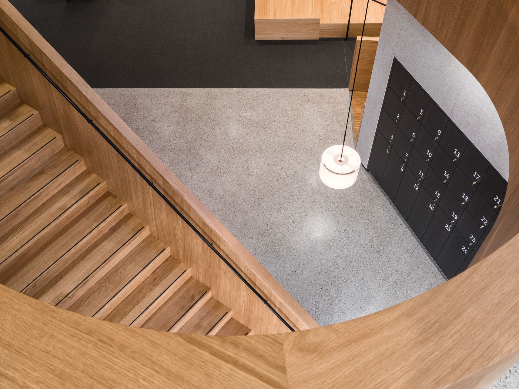 Ein Blick von oben auf eine geschwungene Holztreppe, die in einen Raum mit einer Wand voller nummerierter Schließfächer und einer hängenden Pendelleuchte führt.
