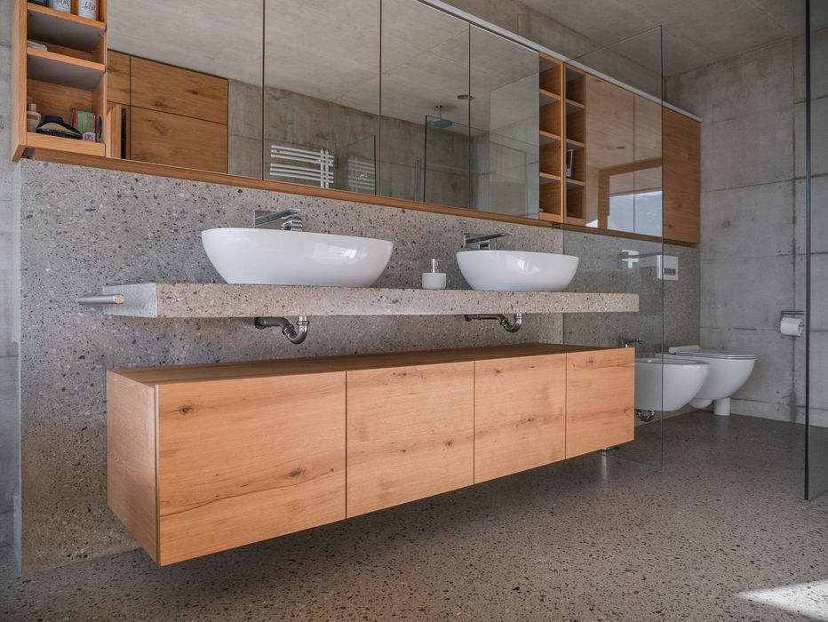 Ein modernes Badezimmer mit einem Doppelwaschtisch auf einer Terrazzo-Platte, hängenden Holzschränken und einer Spiegelfront vor einer Sichtbetonwand.