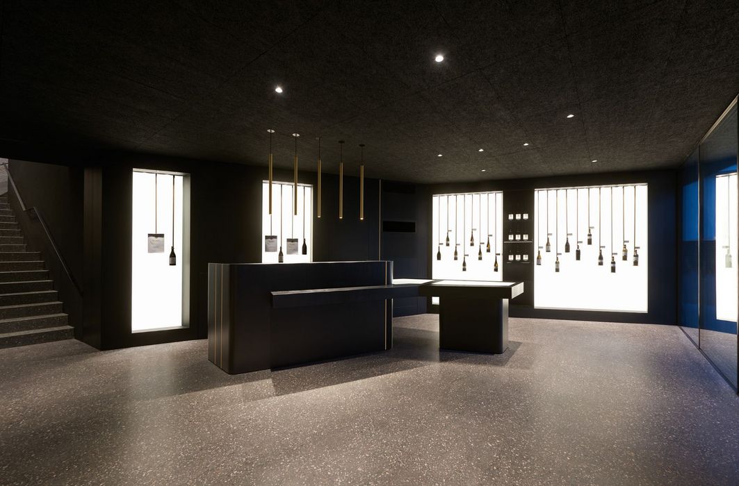 Ein minimalistischer, dunkel gestalteter Verkaufsraum mit einem schwarzen Tresen und von der Decke hängenden Weinflaschen vor hinterleuchteten Fensterfronten.