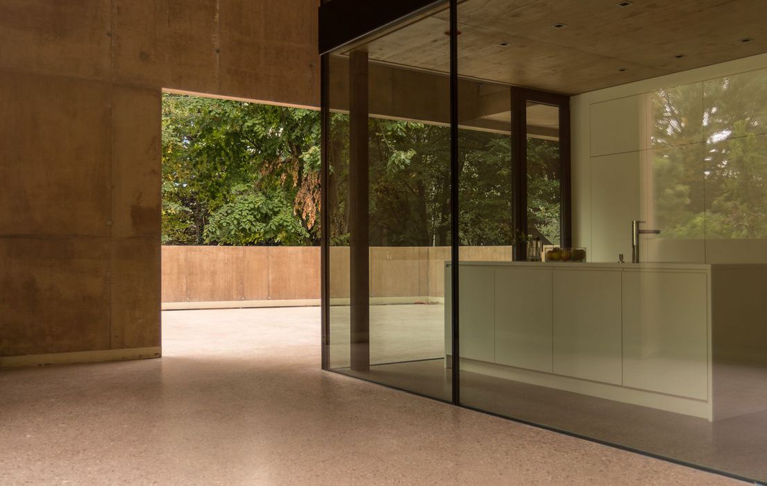 Ein minimalistischer moderner Innenraum mit Betonwänden, der durch eine Glasfront den Blick auf eine Terrasse und das grüne Blätterdach von Bäumen freigibt.