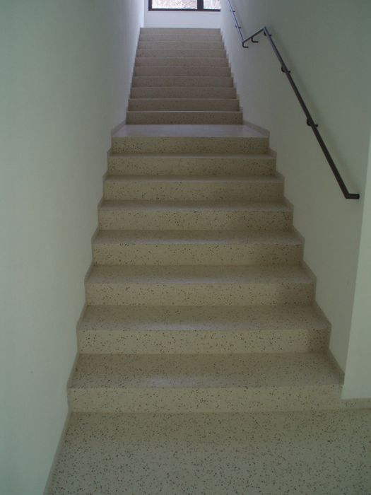 Eine gerade Treppe mit gesprenkelten beigen Stufen führt in einem schlichten, weißen Treppenhaus nach oben zu einem Fenster.