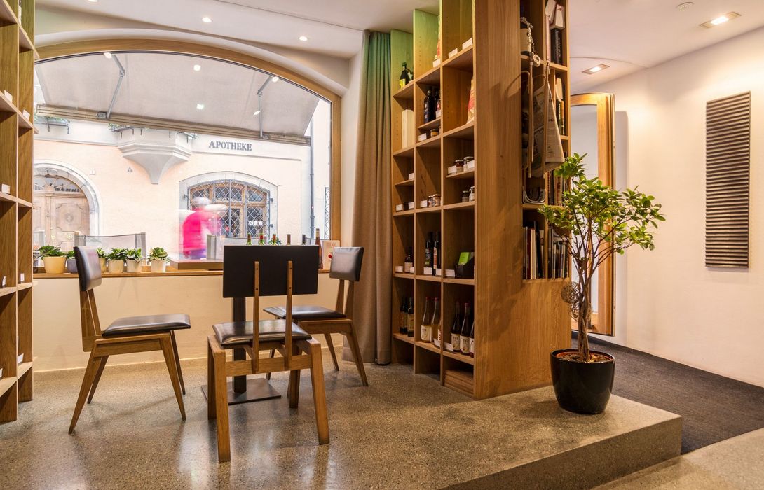 Innenansicht eines Raums mit Holztischen und Stühlen vor einem großen Fenster, das auf eine Straße mit einer Apotheke blickt, daneben ein hohes Holzregal mit Flaschen und eine Zimmerpflanze.