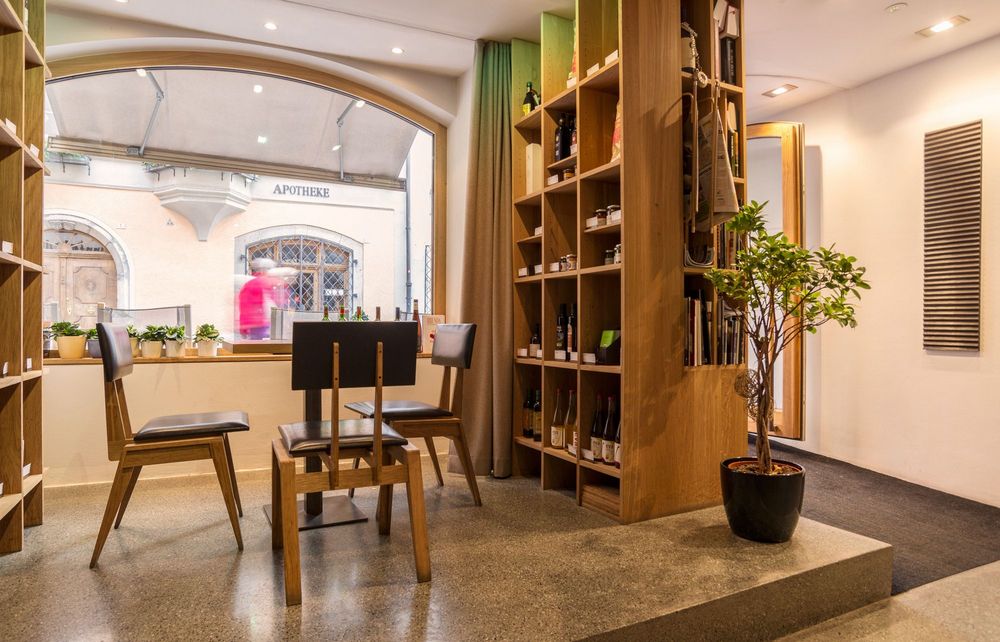 Innenansicht eines Raums mit Holztischen und Stühlen vor einem großen Fenster, das auf eine Straße mit einer Apotheke blickt, daneben ein hohes Holzregal mit Flaschen und eine Zimmerpflanze.