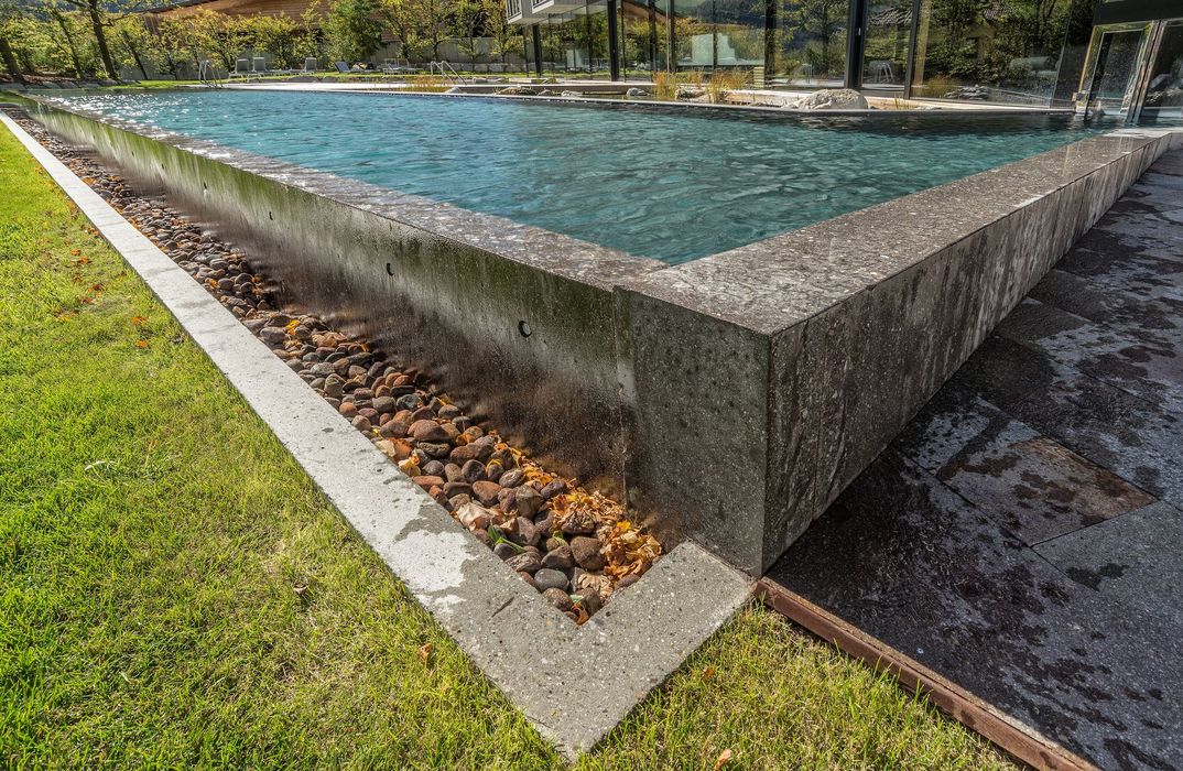 Ein Außenpool mit dunklem Steinrand, der von einer Rasenfläche und einem gepflasterten Bereich umgeben ist, vor einem modernen Gebäude mit Glasfassade.