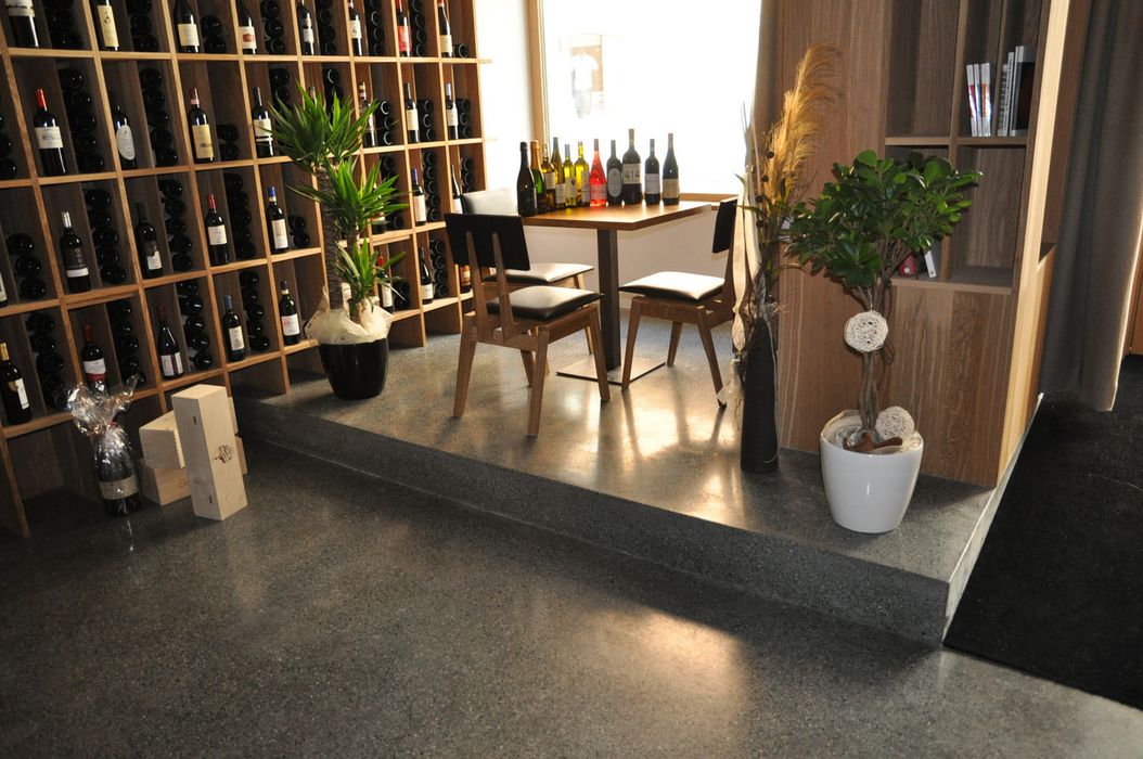 Ein gemütlicher Bereich in einer Weinstube mit einem hölzernen Weinregal, einem kleinen Tisch mit zwei Stühlen und einigen Zierpflanzen vor einem Fenster.