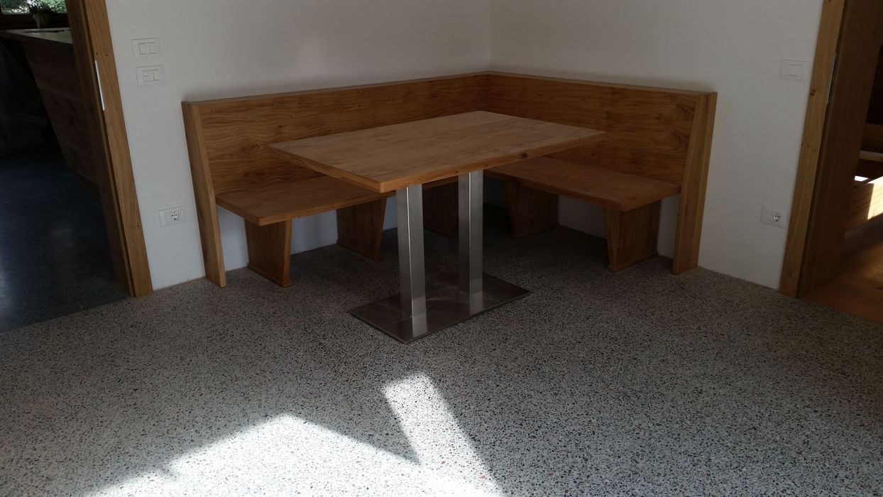 Eine Eckbank aus hellem Massivholz mit einem passenden Holzesstisch auf einem in den Raum ragenden Edelstahlfuß, platziert in einer Ecke vor weißen Wänden und einem Terrazzoboden.