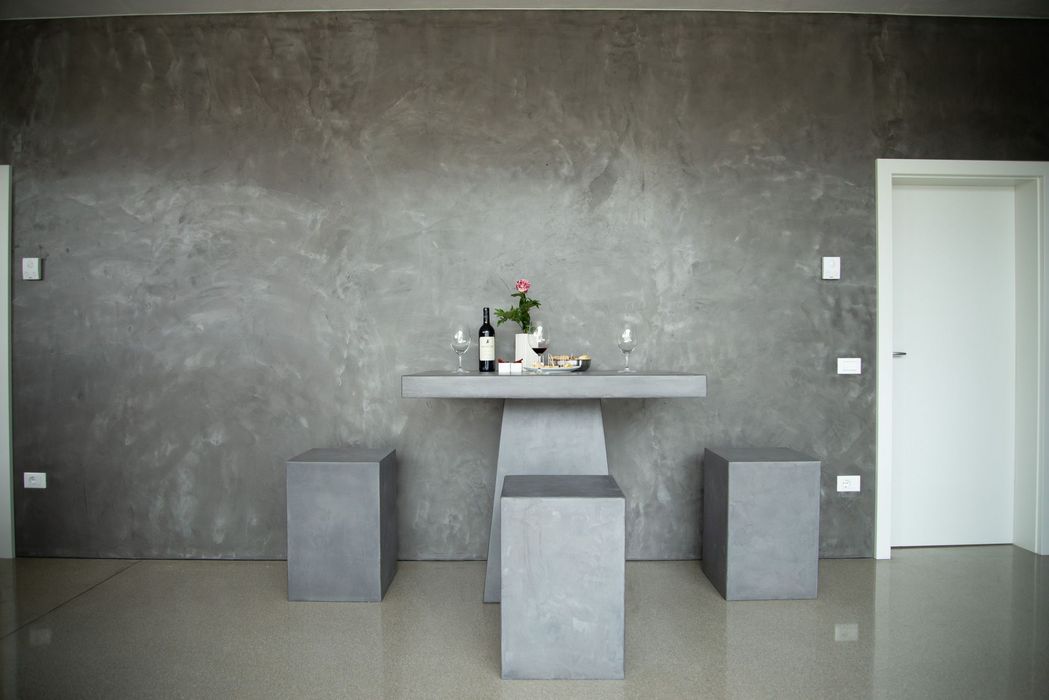Ein minimalistischer Esstisch mit drei passenden Hockern aus grauem Sichtbeton vor einer strukturierten grauen Wand mit einer Flasche Wein und einer Blume in der Mitte.