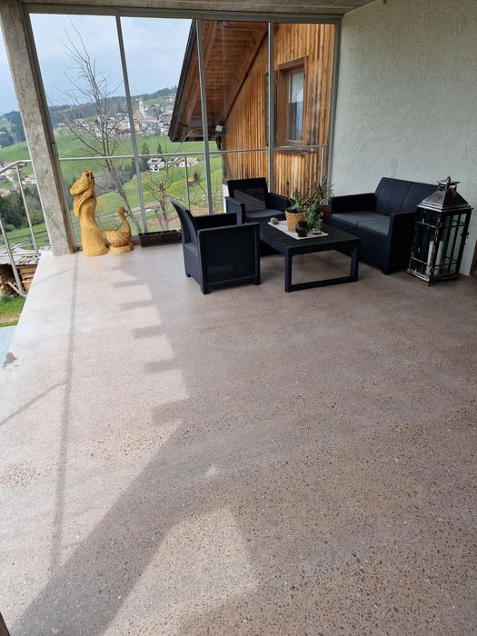 Eine überdachte Terrasse mit schwarzen Rattanmöbeln, bestehend aus zwei Sesseln und einem kleinen Tisch, sowie einer großen Windlicht-Laterne und zwei geschnitzten Holzfiguren vor einer verglasten Front mit Blick auf eine hügelige Alpenlandschaft.