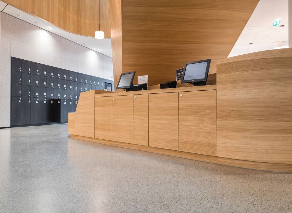 Ein hölzerner Empfangstresen mit zwei digitalen Kassenbildschirmen steht vor einer grauen Wand mit nummerierten Schließfächern in einem modernen Innenraum.