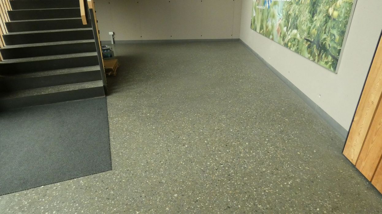 Innenraum mit geflecktem Steinfußboden, einer grauen Treppe links, einer grauen Wand mit einem grünen Wandbild rechts und einem kleinen Stapel flacher Kartons in der Ecke.