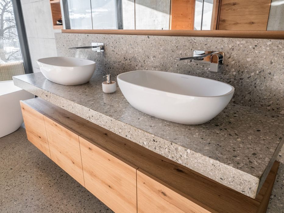 Ein modernes Doppelwaschbecken mit zwei weißen Aufsatzwaschbecken auf einer grauen Steinplatte, kombiniert mit einem hölzernen Unterschrank und Wandarmaturen.