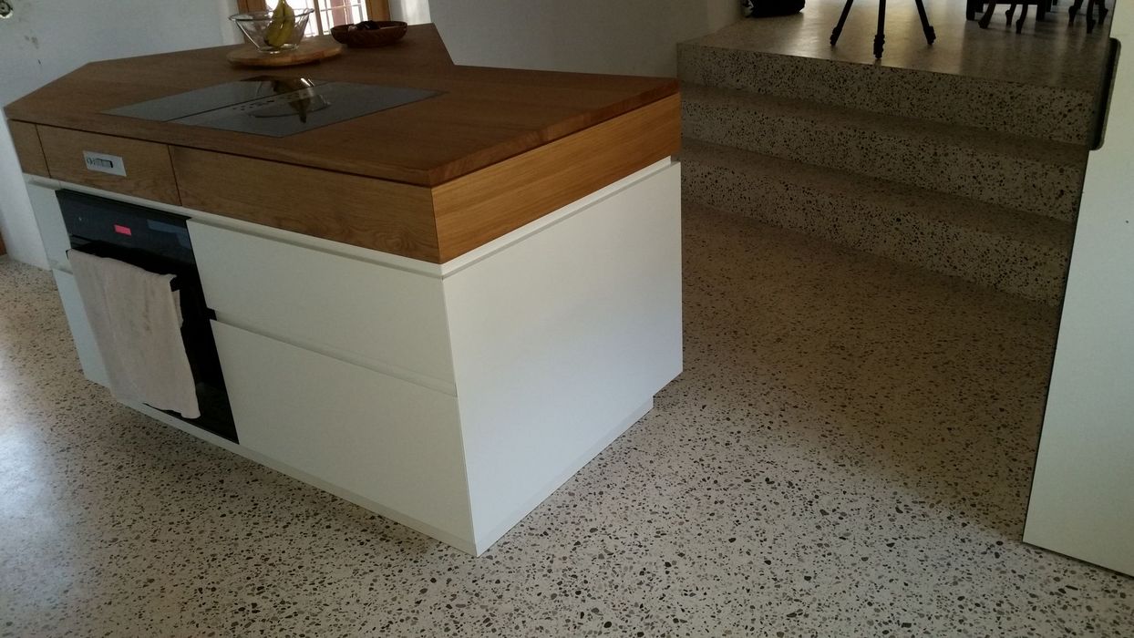 Eine Kücheninsel mit weißer Front und Holzarbeitsplatte steht auf einem Terrazzoboden vor einer kleinen Treppe.