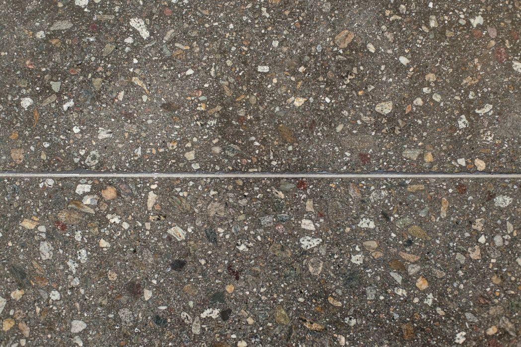 Nahaufnahme einer texturierten Betonoberfläche mit eingelassenen, verschiedenfarbigen Kieselsteinen, die durch eine horizontale Fuge in zwei Hälften geteilt wird.