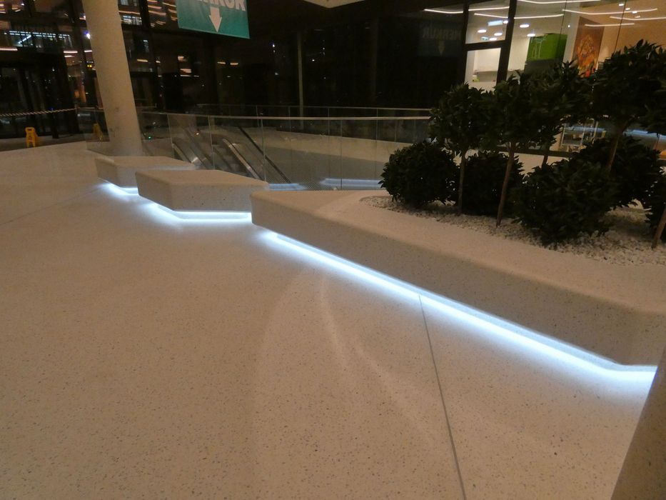 Moderne Sitzgelegenheiten aus hellem Stein mit indirekter LED-Unterbodenbeleuchtung in einem sauberen Einkaufszentrum vor einer Glasbrüstung mit Rolltreppe.