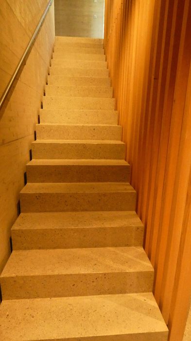 Eine gerade Treppe aus hellem Stein führt nach oben, flankiert von einer Wand aus Holzlatten auf der rechten Seite und einer glatten Wand mit Handlauf auf der linken Seite.