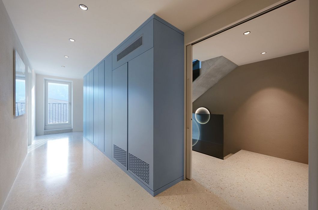 Ein heller Innenraum mit einem zentralen, blauen Einbauschrank, der den Flur von einem Treppenhaus abgrenzt.