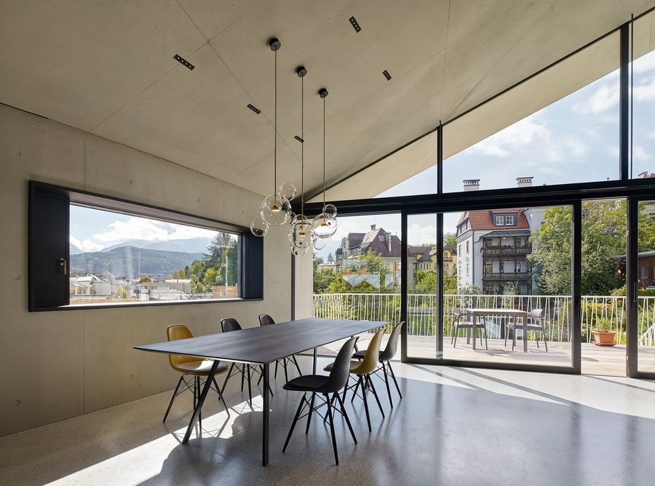 Ein minimalistisch eingerichtetes Esszimmer mit großem dunklen Tisch, Stühlen in Gelb und Anthrazit sowie einer markanten Glas-Pendelleuchte, gelegen in einem Haus mit Sichtbetonwänden, großen Panoramafenstern und Blick auf eine Terrasse und Nachbargebäude.