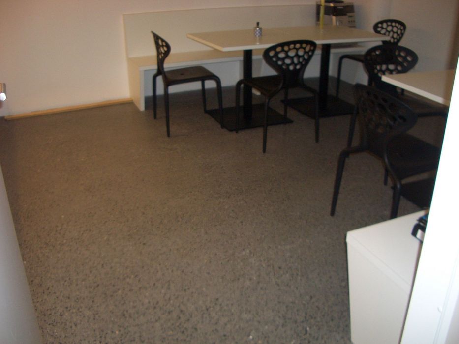 Ein minimalistischer Essbereich mit einem weißen Tisch, einer weißen Sitzbank und mehreren schwarzen Stühlen mit organischem Lochmuster auf einem grauen Boden.