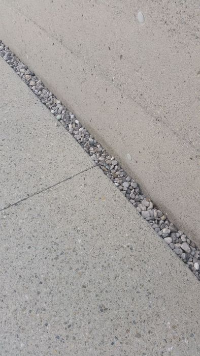 Eine graue Betonmauer, die an eine gepflasterte Fläche grenzt, wobei ein schmaler Streifen aus kleinen Kieselsteinen die Trennung zwischen Wand und Boden markiert.