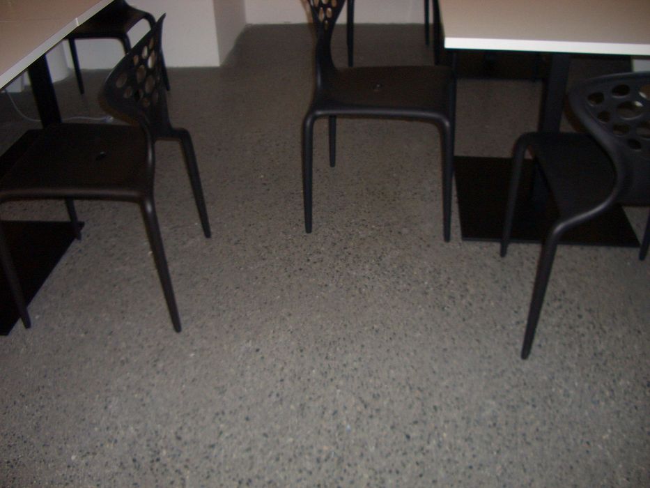Blick auf einen grauen, gesprenkelten Boden mit mehreren schwarzen Stühlen, die an weißen Tischen stehen.