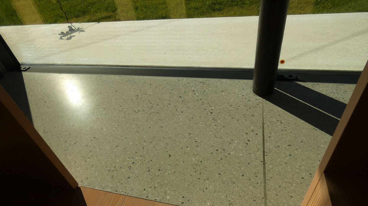 Ein Blick durch eine bodentiefe Glasfront auf einen Terrazzoboden im Innenraum und eine angrenzende helle Terrasse im Außenbereich.