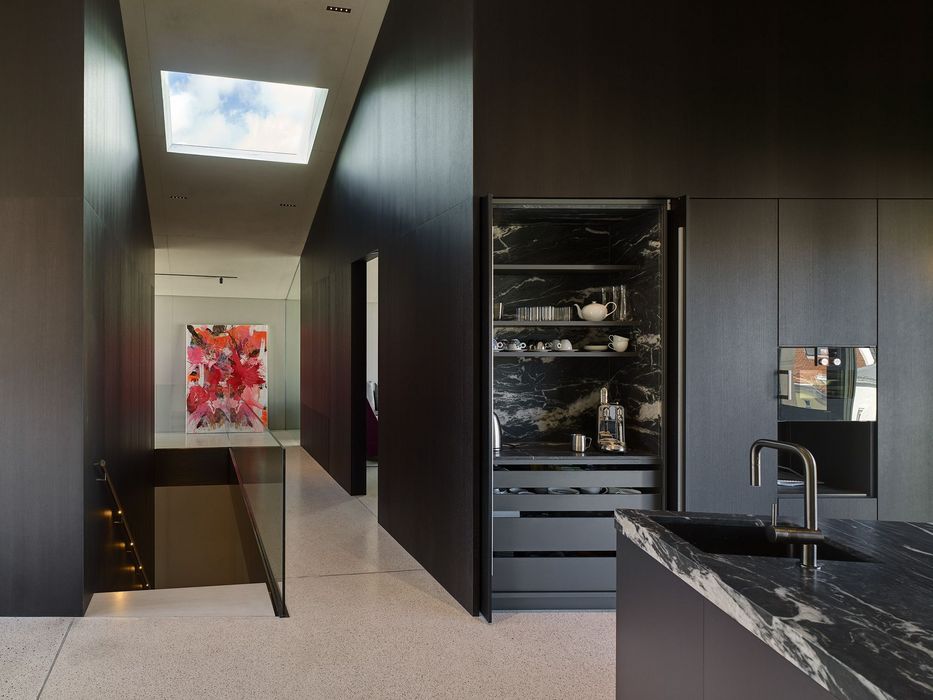 Ein minimalistisch gestalteter Innenraum mit einer dunklen Holzwand, einem offenen Küchenschrank aus schwarzem Marmor und Treppenabgang, ergänzt durch ein abstraktes rotes Kunstwerk am Ende des Flurs unter einem Dachfenster.