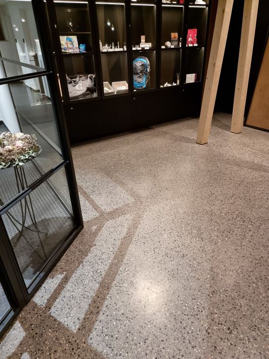 Ein moderner Verkaufsbereich mit Terrazzoboden, einer schwarzen Glasvitrine mit verschiedenen Produkten, einem Rucksack und zwei Holzstützen in einem minimalistischen Interieur.
