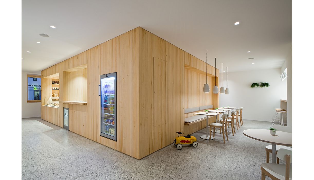 Innenansicht eines modernen Cafés mit hellen Holzmöbeln, Sitzbänken, kleinen Tischen und einer Vitrine mit Getränken sowie einem Spielzeugauto am Boden.