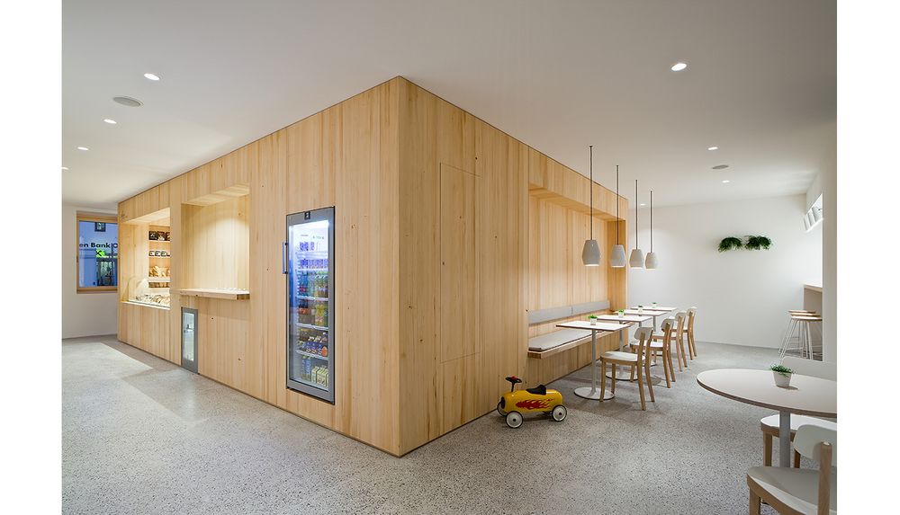 Innenansicht eines modernen Cafés mit hellen Holzmöbeln, Sitzbänken, kleinen Tischen und einer Vitrine mit Getränken sowie einem Spielzeugauto am Boden.