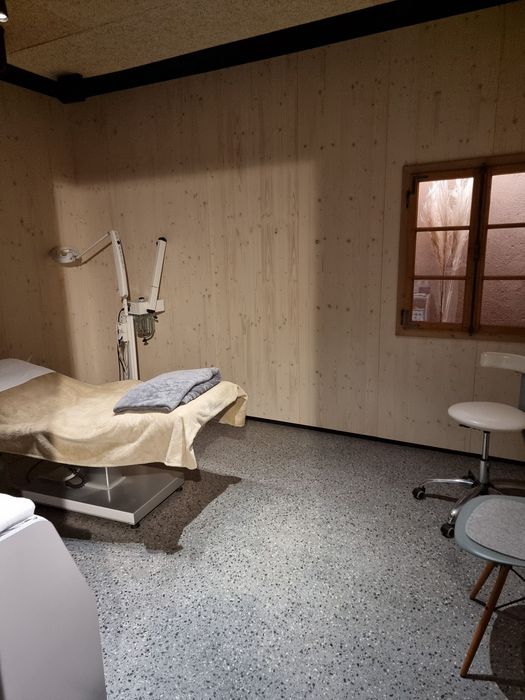 Ein minimalistischer Behandlungsraum mit einer Untersuchungsliege, einem kosmetischen Bedampfer, einem Hocker sowie holzverkleideten Wänden und einem kleinen Fenster.
