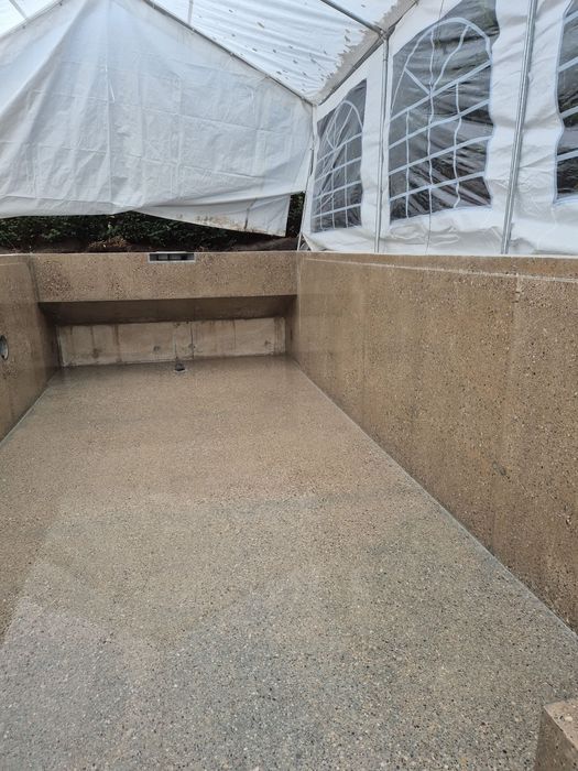 Ein leeres Schwimmbecken aus beigem Sichtbeton unter einem provisorischen weißen Zeltdach.