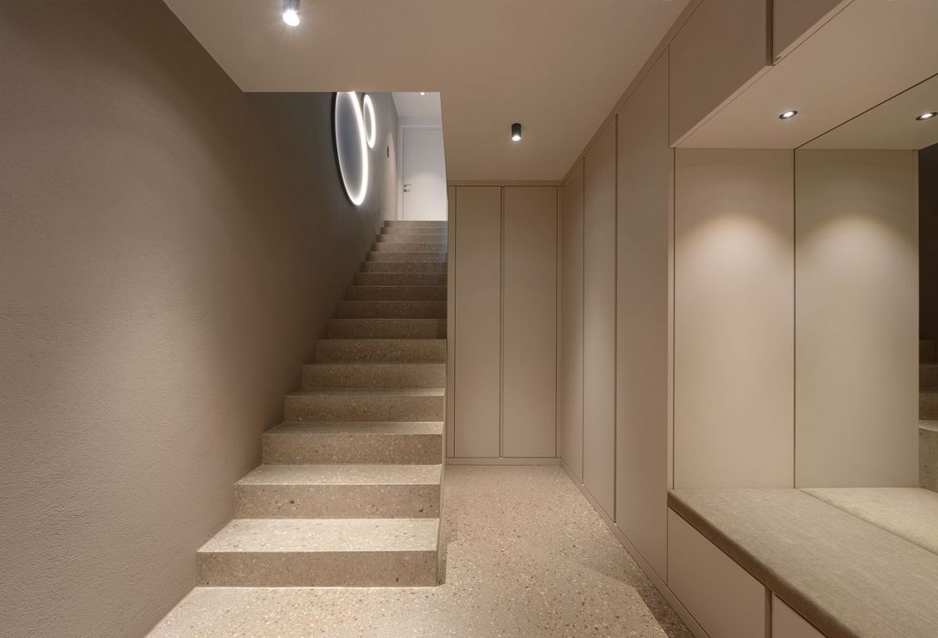 Ein minimalistischer Flur mit einer Steintreppe, die in einen beleuchteten Durchgang führt, flankiert von einer glatten hellen Wand und Einbauschränken.