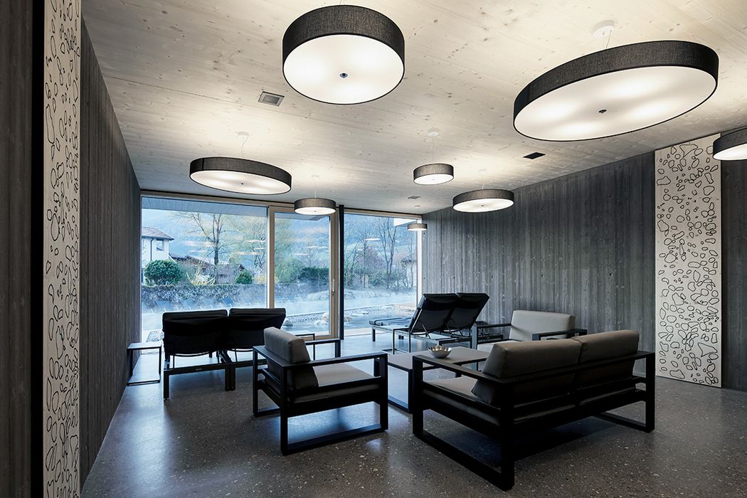 Ein moderner, minimalistischer Ruheraum mit grauen Wänden, schwarzen Loungesesseln und runden Deckenleuchten, mit Blick durch eine Fensterfront auf eine Außenanlage.