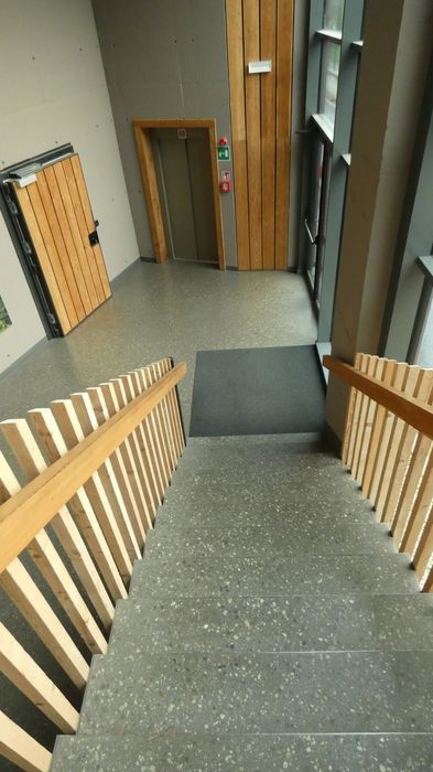 Blick von einer Treppe mit Holzgeländer auf das Treppenpodest eines modernen Gebäudes mit Betonwänden, einem Aufzug und einer verglasten Außenwand.