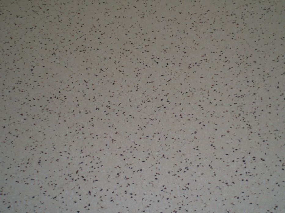 Nahaufnahme einer hellbeige gesprenkelten Kunststoffoberfläche mit unregelmäßigen dunklen Einschlüssen.